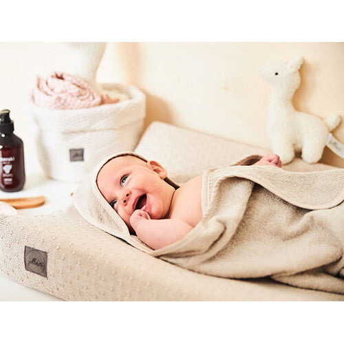 Een lachende baby ligt gewikkeld in een Jollein badcape in nougat kleur op een aankleedkussen.