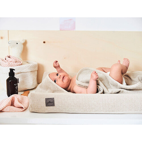 Een baby ligt op de rug op een aankleedkussen, deels bedekt met een Jollein badcape in nougat kleur.