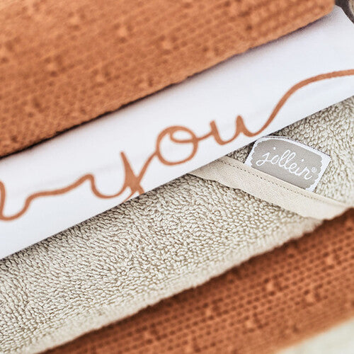 Detail van een Jollein badcape in nougat kleur met het merklabel, liggend in een stapel textiel.