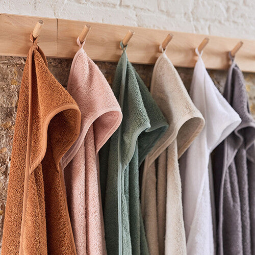 Een rij Jollein badcapes in verschillende kleuren hangt aan houten knoppen tegen een bakstenen muur.