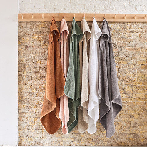 Een rij Jollein badcapes in verschillende kleuren hangt aan houten knoppen tegen een bakstenen muur.