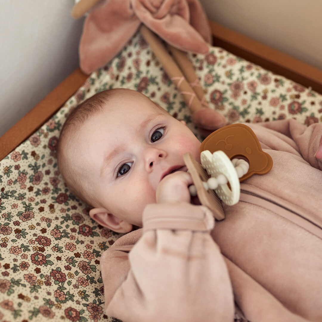 Een baby ligt op een aankleedkussen en heeft het bijtspeelgoed met de bruine teddybeer vorm in de mond.
