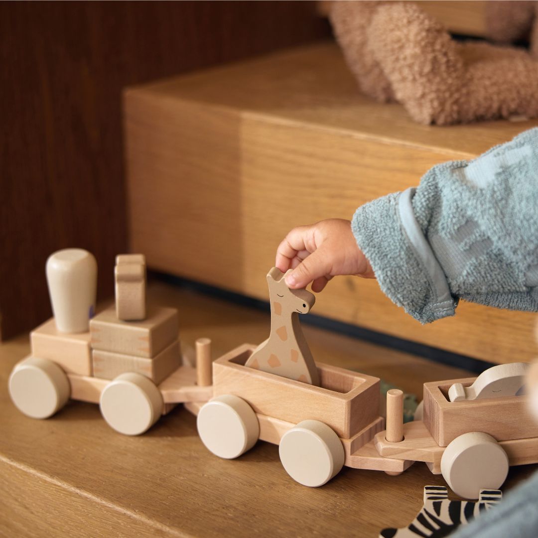 Een kinderhand in een blauwe badjas speelt met de Jollein houten blokkentrein en plaatst een giraffe in een wagon.
