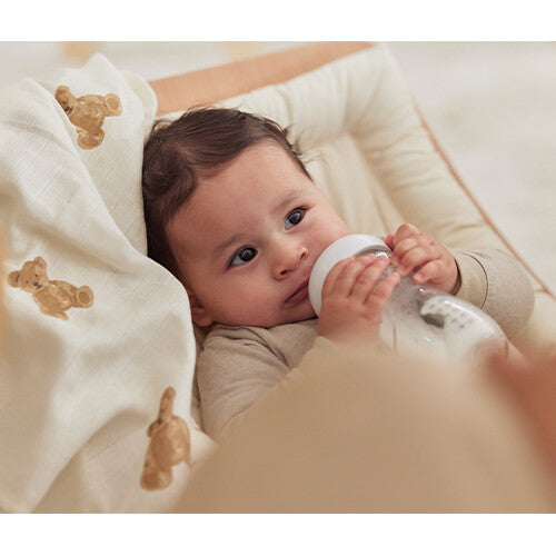 Een baby drinkt uit een flesje, liggend naast een Jollein hydrofiele doek met teddybeer patroon.