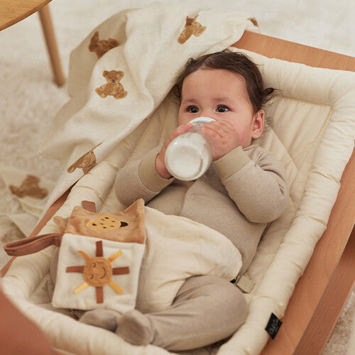 Een baby drinkt uit een flesje in een wiegje, met een Jollein hydrofiele doek met teddybeer patroon ernaast.