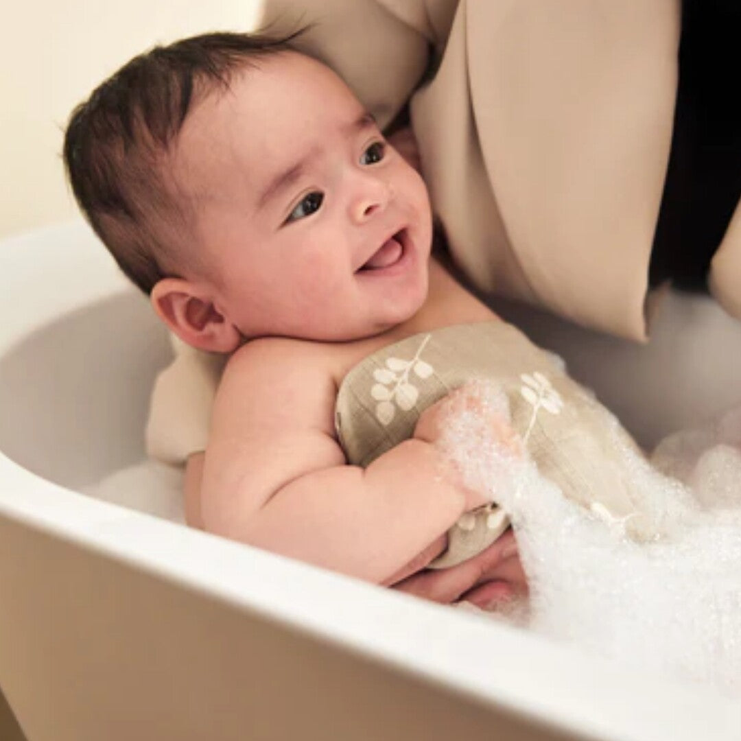 Een lachende baby in bad met een Jollein hydrofiel washandje met bladpatroon en bubbels.