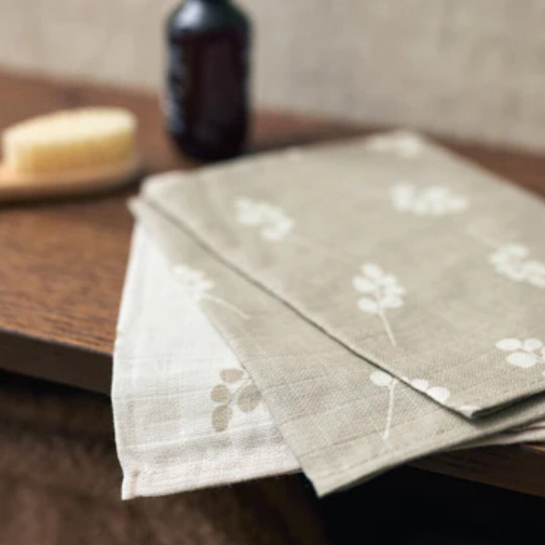 Twee Jollein hydrofiele washandjes met bladpatroon op een houten plank, met badaccessoires op de achtergrond.