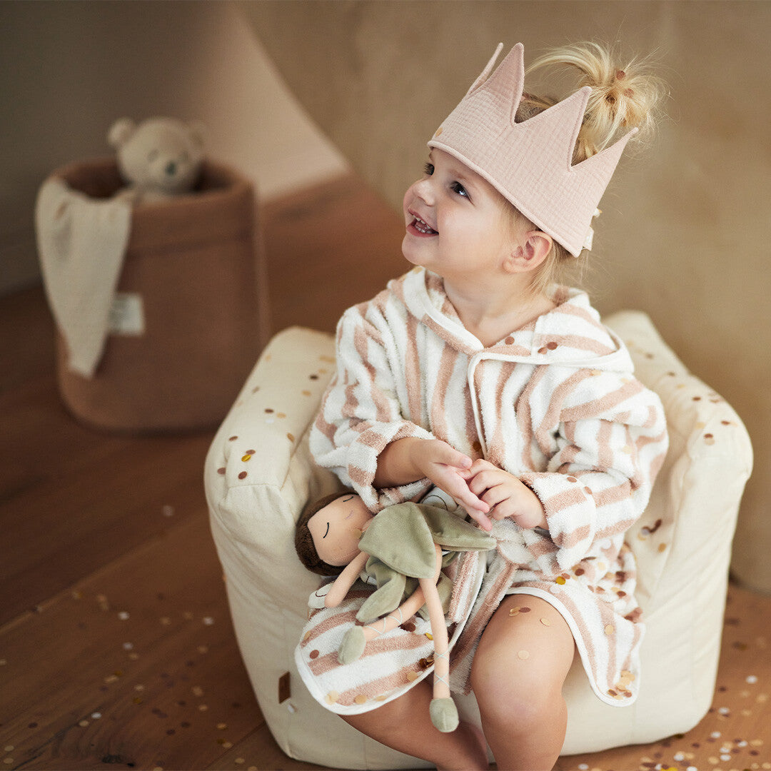 Een kind met roze kroon en badjas zit in de Jollein kinderfauteuil en houdt een pop vast.