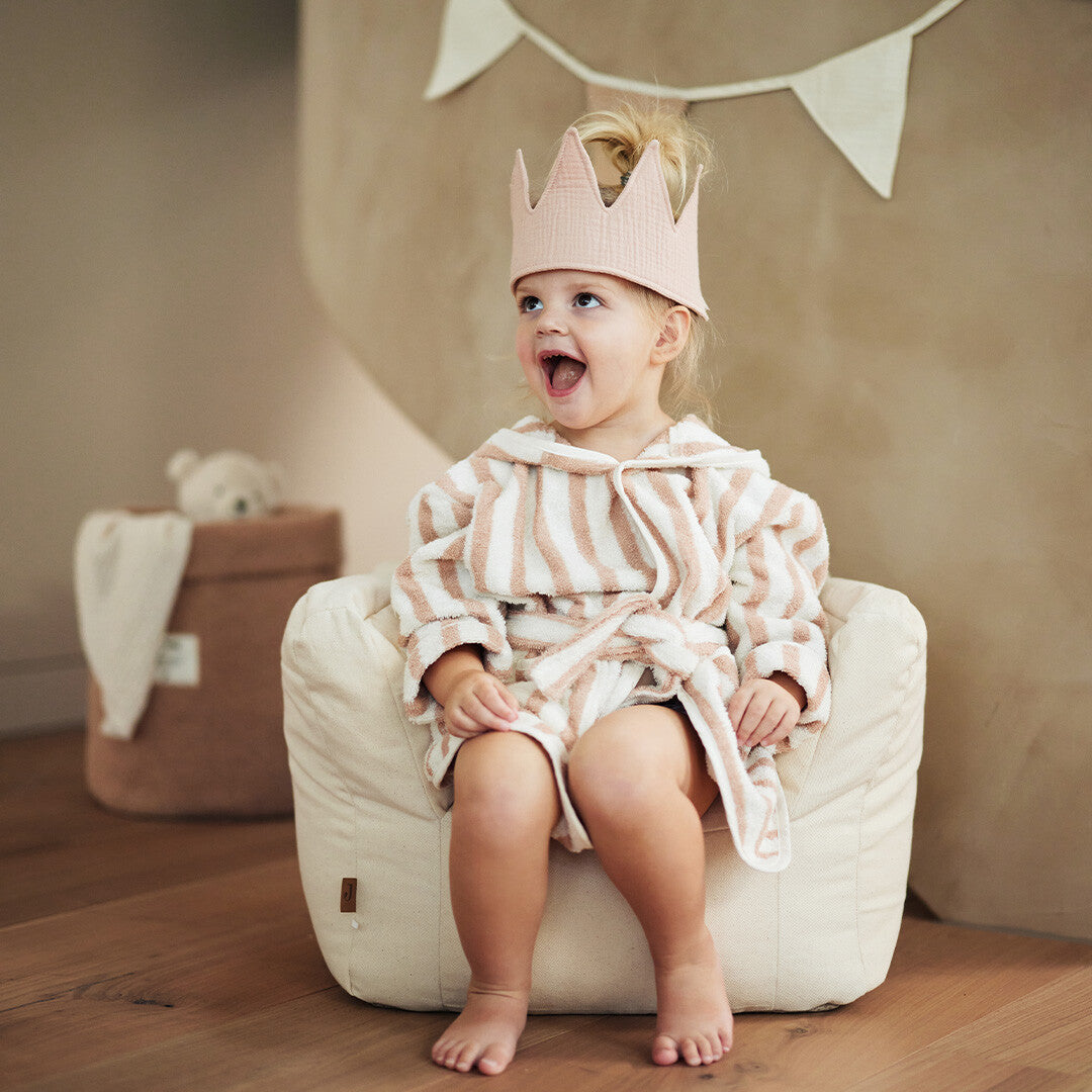 Een kind met roze kroon en badjas zit lachend in de Jollein kinderfauteuil.