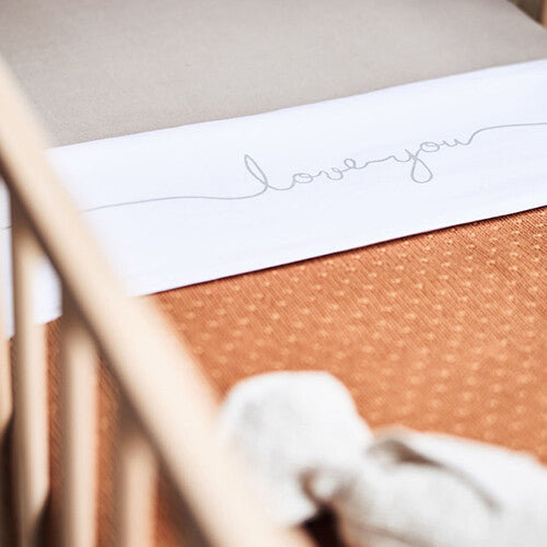 Detail van een wit laken met 'love you' erop, liggend in een ledikant met een bruine deken.