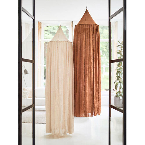 Twee Jollein klamboes, een beige en een bruine, hangen in een kamer, gezien door een deuropening.