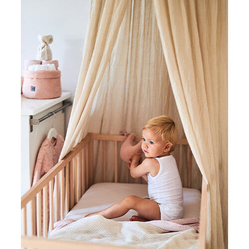 Een baby zit in een houten ledikant onder een Jollein beige bedhemel.