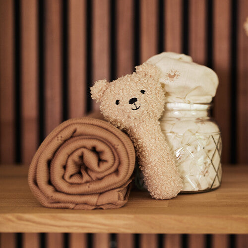 De Jollein knijprammelaar teddybeer ligt naast een opgerolde deken en een pot met watten op een houten plank.