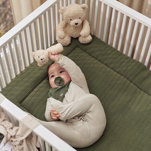 Een baby in een ledikant met groene mat reikt naar de Jollein knijprammelaar teddybeer.