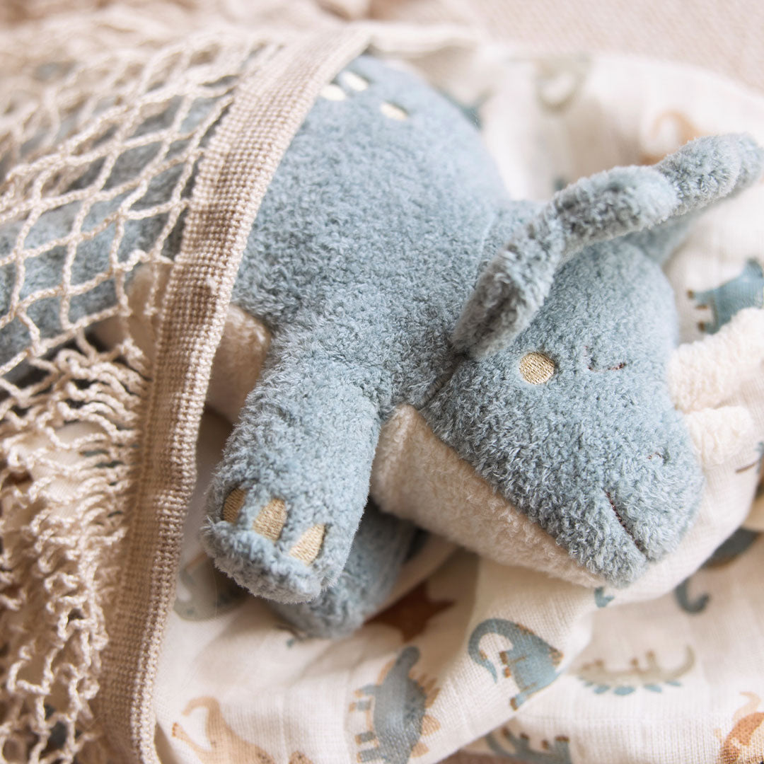De Jollein pluchen triceratops knuffeldino ligt op een deken met dinoprint, deels onder een netje, vanuit een andere hoek.