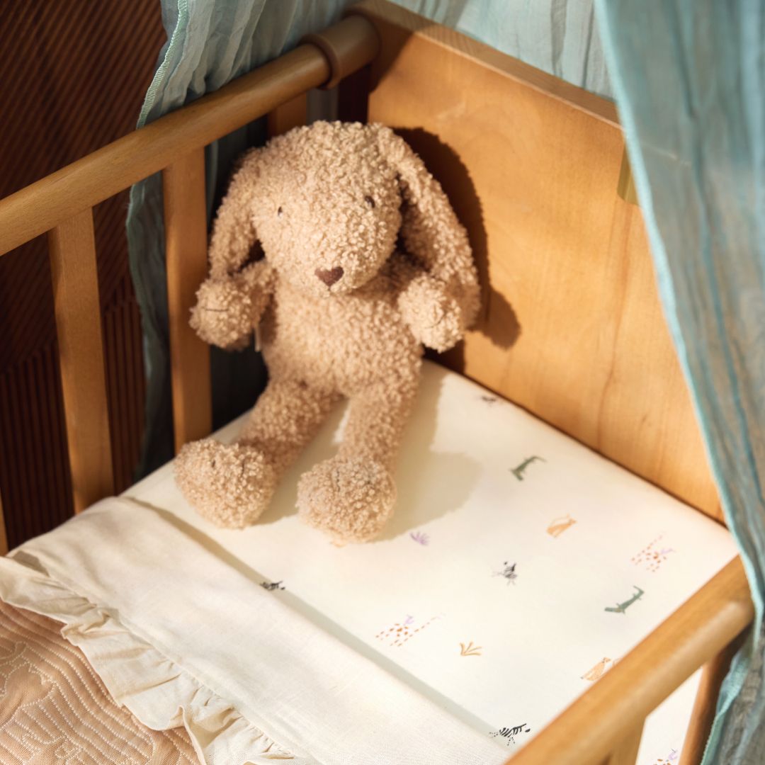 Jollein pluchen knuffelkonijn zit in een houten babywiegje met een wit laken.