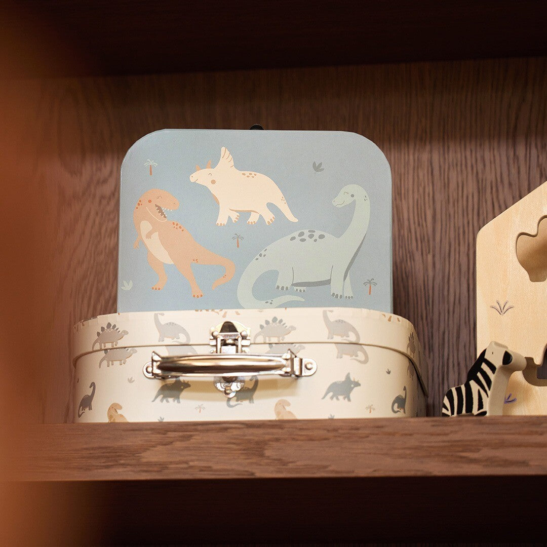 De Jollein kinderkoffers met dinosauruspatroon staan op een houten plank, met de blauwe koffer bovenop de beige.