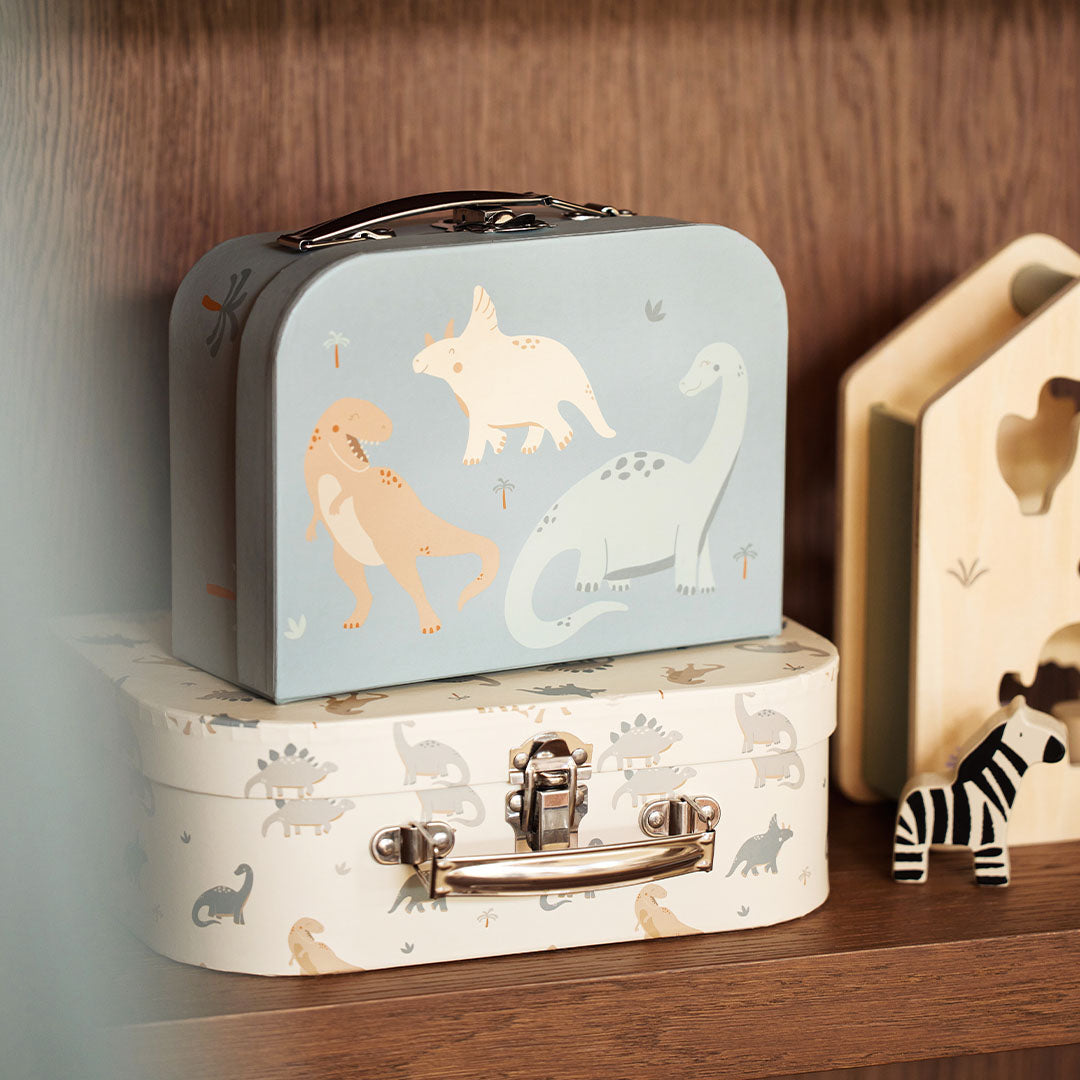 De Jollein kinderkoffers met dinosauruspatroon staan op een houten plank, met de blauwe koffer bovenop de beige.