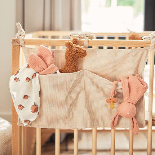 Jollein baby organizer van nougat corduroy hangend aan een houten ledikant, gevuld met babyspeelgoed en een speen.