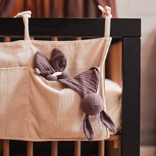 Jollein baby organizer van nougat corduroy hangend aan een donkerhouten ledikant, gevuld met een paars knuffeldoekje.