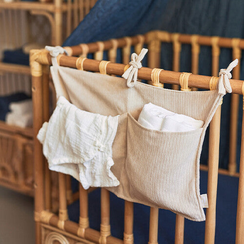 Jollein baby organizer van nougat corduroy hangend aan een rotan ledikant, gevuld met witte babykleding.