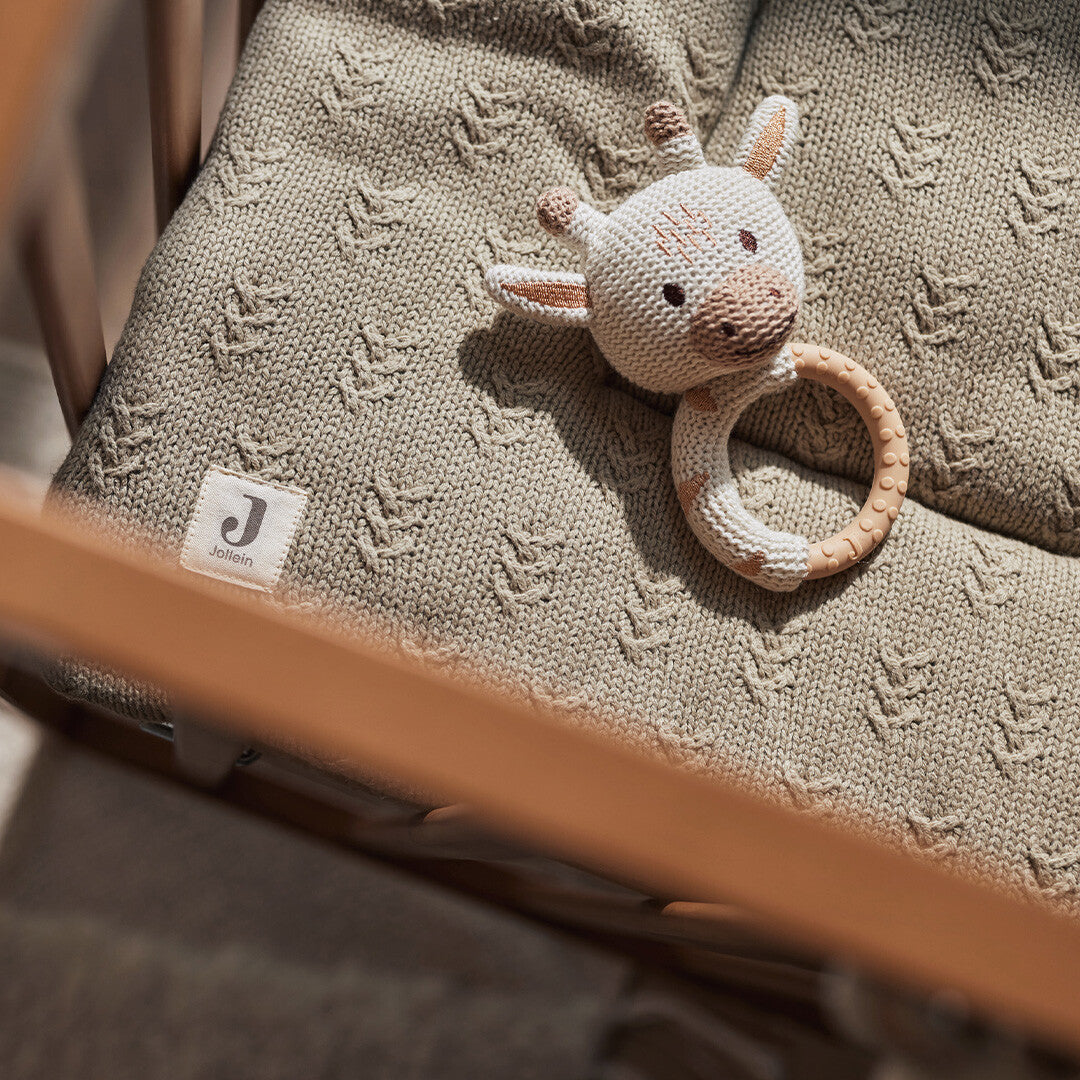 Jollein babyspeelgoed rammelaar met bijtring in de vorm van een giraffe, liggend op een gebreide deken in een wieg.