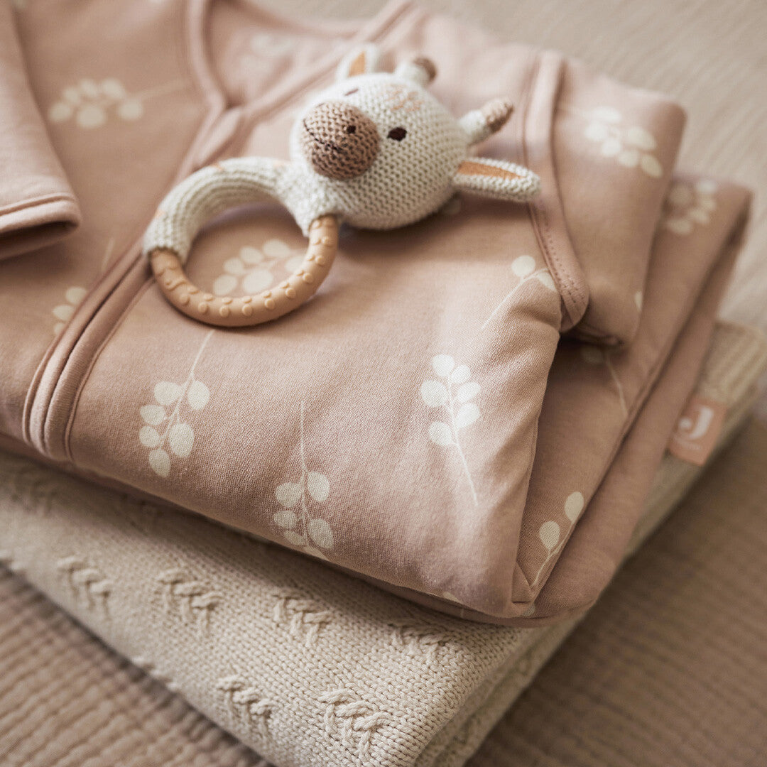 Jollein babyspeelgoed rammelaar met bijtring in de vorm van een giraffe, liggend op een stapel roze babykleding en een deken.