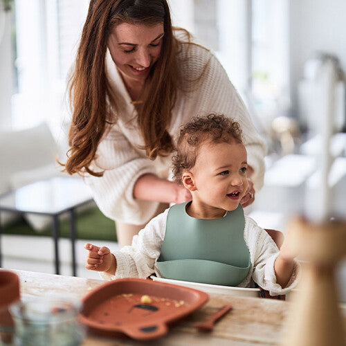 Een baby draagt het Jollein siliconen slabbetje tijdens het eten, met een volwassene erachter.