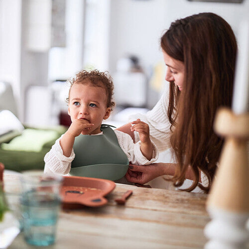 Een baby met het Jollein siliconen slabbetje aan tafel, terwijl een volwassene de sluiting aanpast.