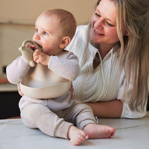 Baby draagt een Jollein beige siliconen slabbetje en kauwt op een bijtring, met een lachende vrouw op de achtergrond.