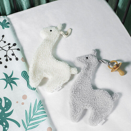 Twee Jollein pluchen lama speendoeken, één off-white en één grijs, liggen op een wit babybedje.