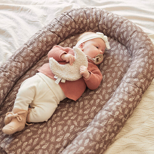 Een baby ligt in een babynest en houdt de Jollein maan speendoek vast.
