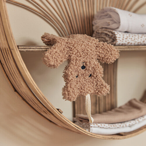 Het Jollein teddy bear speendoekje hangt aan een rotan plankje, met opgevouwen babyartikelen op de achtergrond.