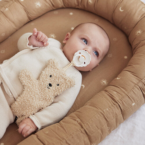 Een baby met een speen en de jollein teddybeer speendoek ligt in een bruin babynestje.
