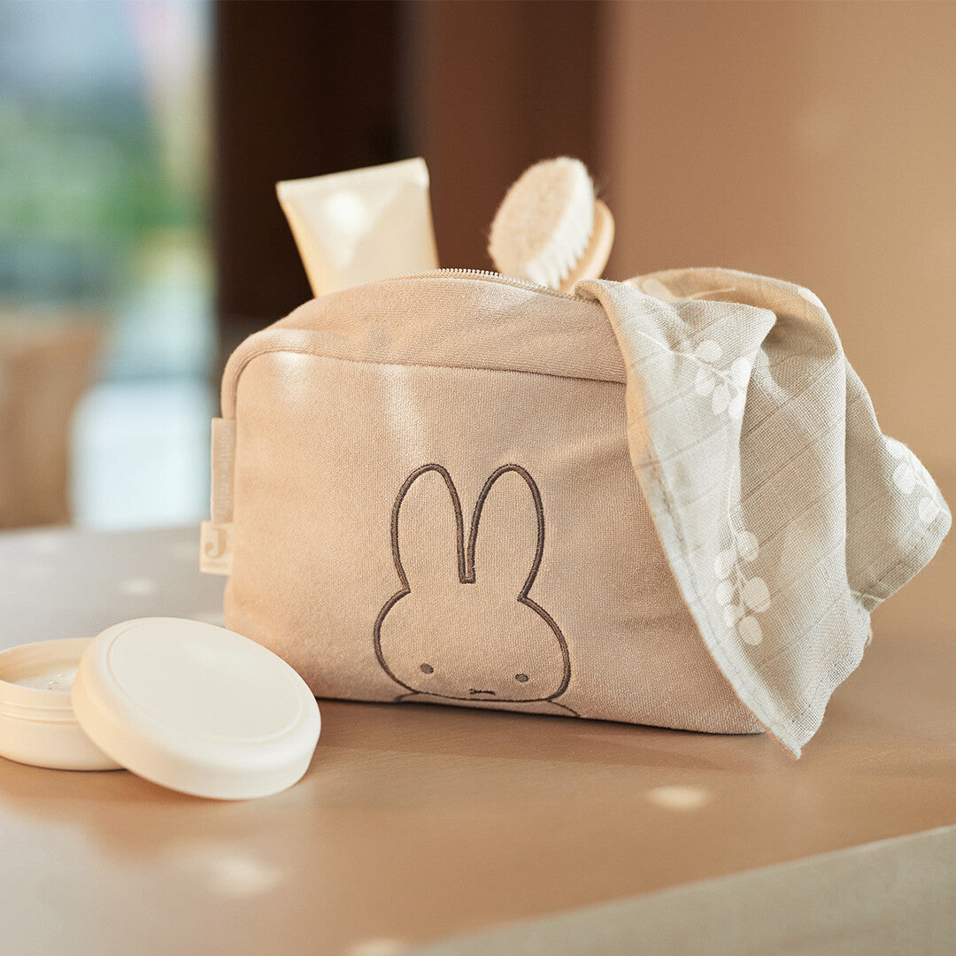 Jollein beige badstof toilettas met Miffy, gevuld met babyverzorgingsproducten op een houten ondergrond.
