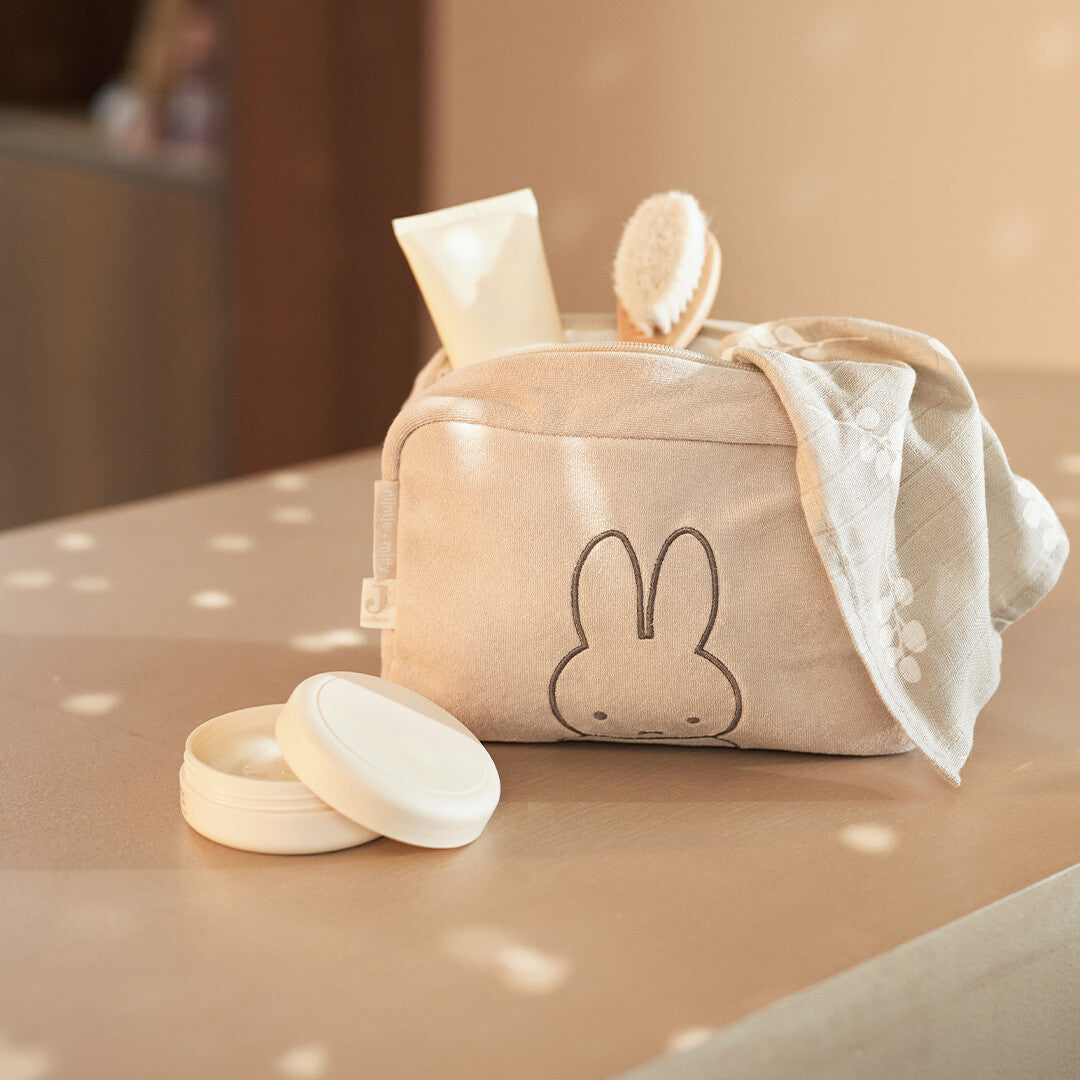Jollein beige badstof toilettas met Miffy, gevuld met babyverzorgingsproducten en een doek ernaast.