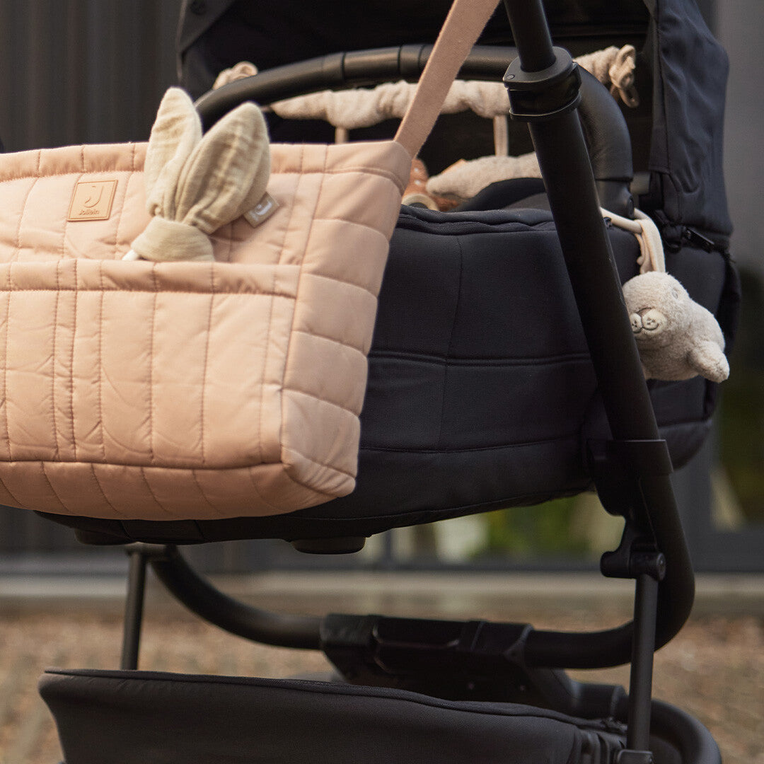 Jollein wagenhanger met pluche zeehond aan een zwarte kinderwagen, naast een roze luiertas met konijnenoren.