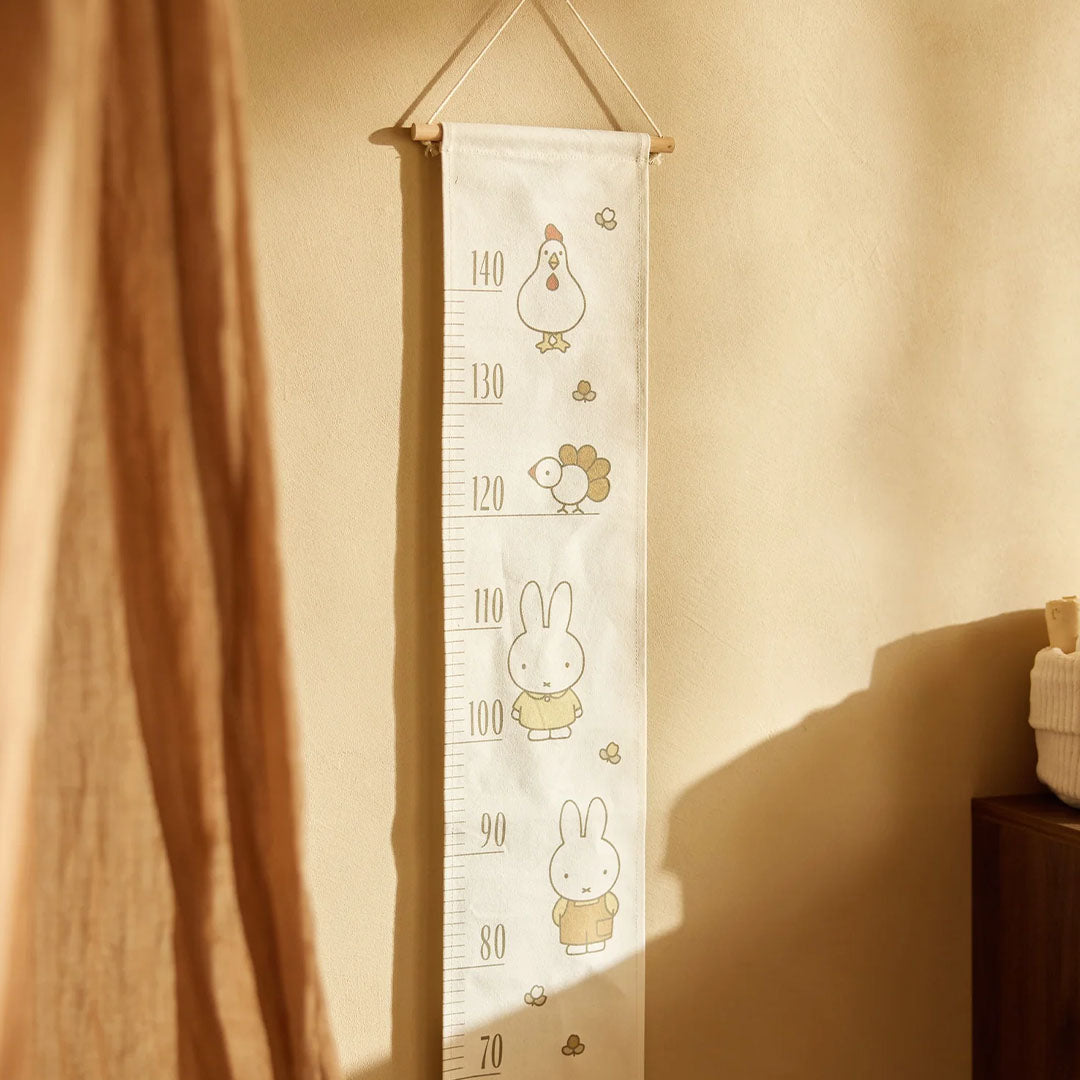 Beige groeimeter met Miffy en boerderijdieren hangt aan een lichte muur naast een bruin gordijn.