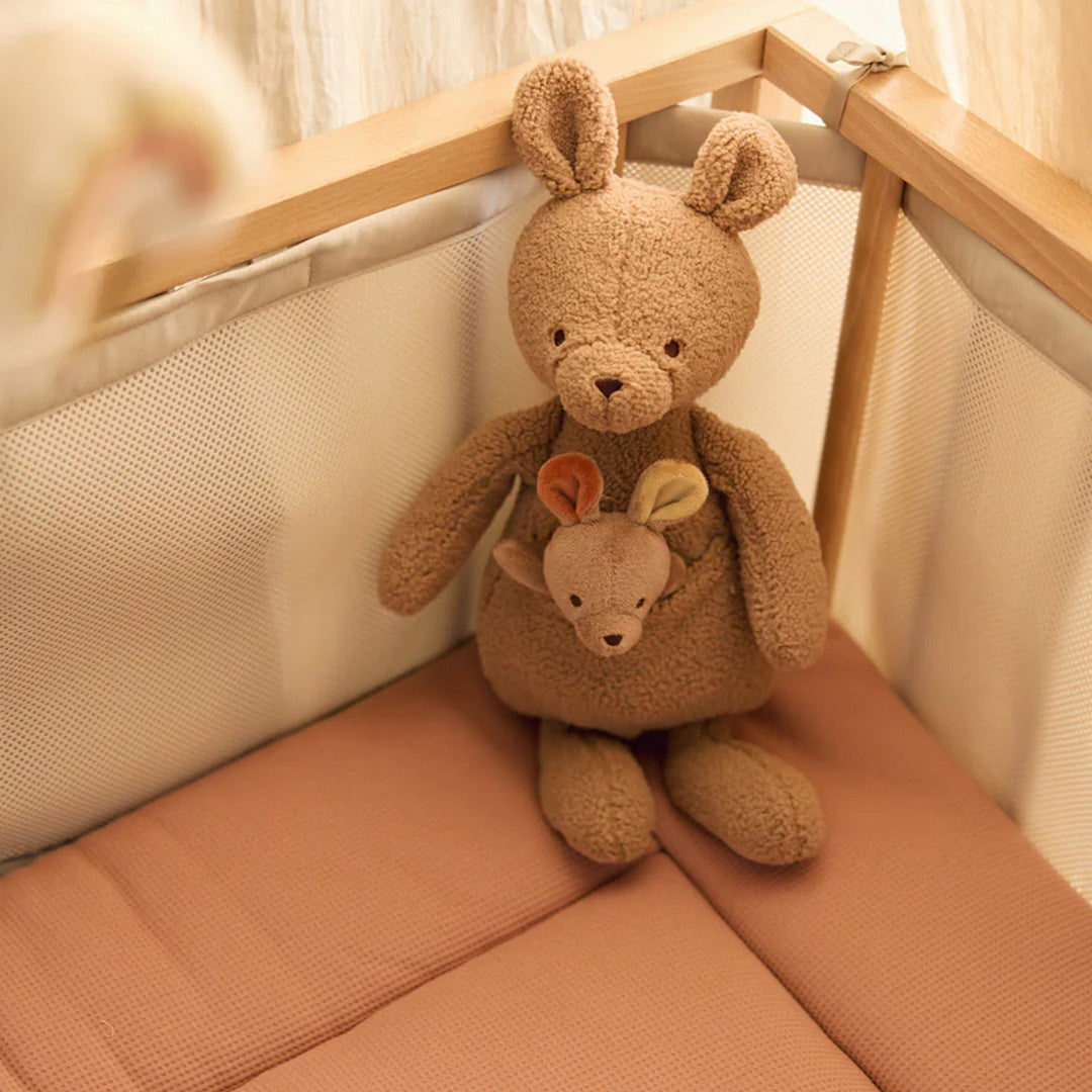 Jollein pluchen knuffelkangoeroe met baby in de buidel, zittend in een babybedje met roze matras.