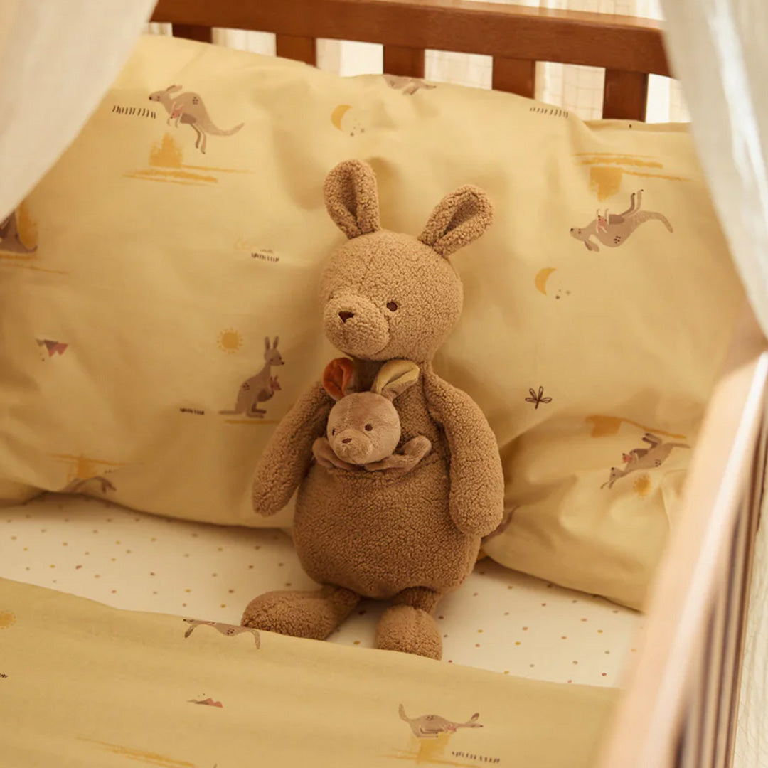 Jollein pluchen knuffelkangoeroe met baby in de buidel, zittend in een babybedje met geel beddengoed.