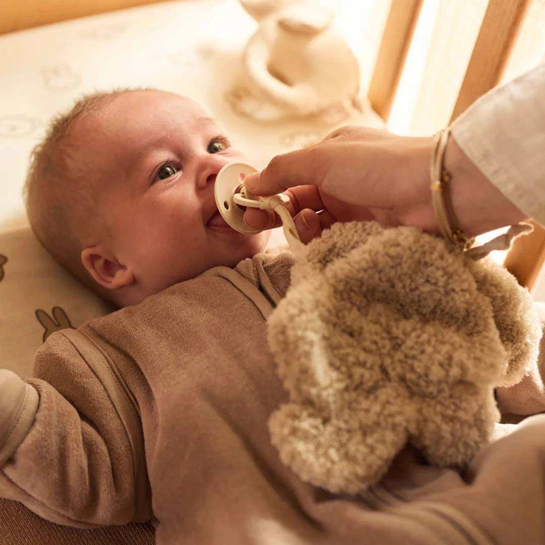 Een baby in een wieg lacht terwijl een hand de Jollein olifant speendoek bij de speen houdt.