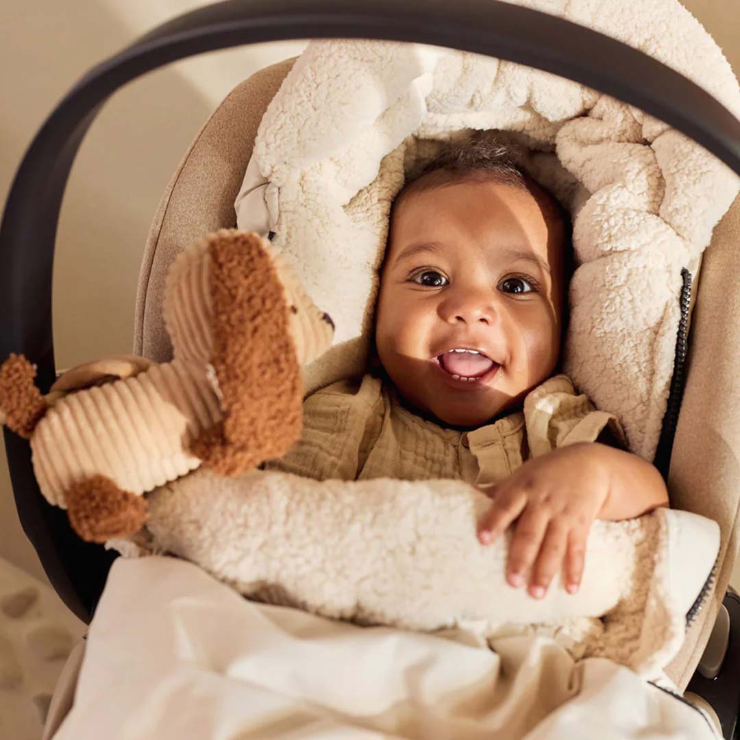 Een lachende baby ligt in een autostoel en speelt met de Jollein pluche honden wagenhanger.