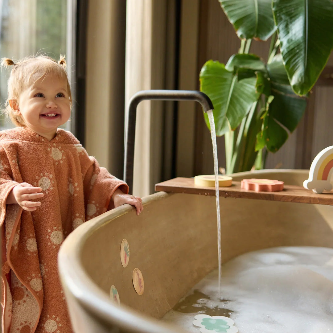 Een lachende baby in een oranje badponcho kijkt naar het bad met Jollein badpuzzelstukken op een houten plank.