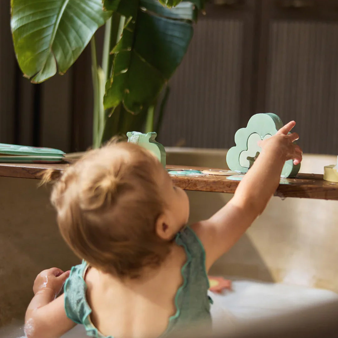Een baby in bad reikt naar een groen wolk-vormig Jollein badpuzzelstuk op een houten plank.