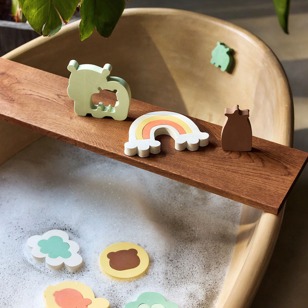 Jollein badpuzzelstukken in de vorm van een kangoeroe, regenboog en flesje staan op een houten plank over een bad met schuim.
