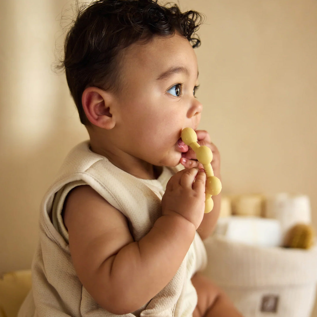 Een baby met krullen houdt de beige Jollein bijtring vast en kijkt opzij.