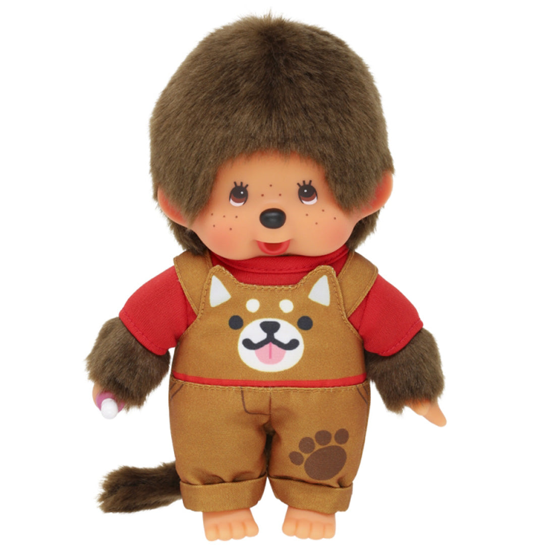 Monchhichi pop jongen met bruine vacht, rode trui en bruine tuinbroek met hondenprint, met een speen in de hand.