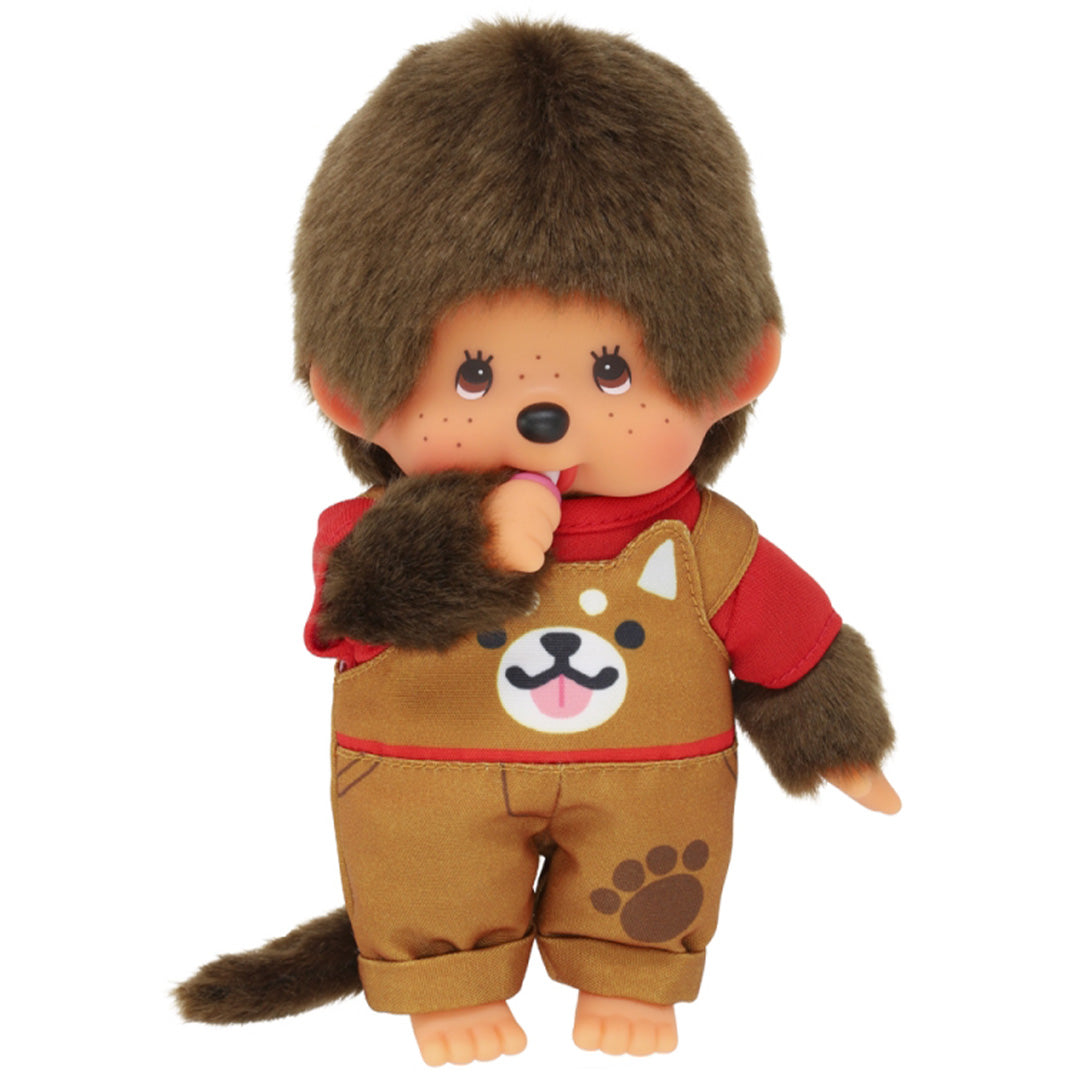 Monchhichi pop jongen met bruine vacht, rode trui en bruine tuinbroek met hondenprint, met een speen aan de mond.