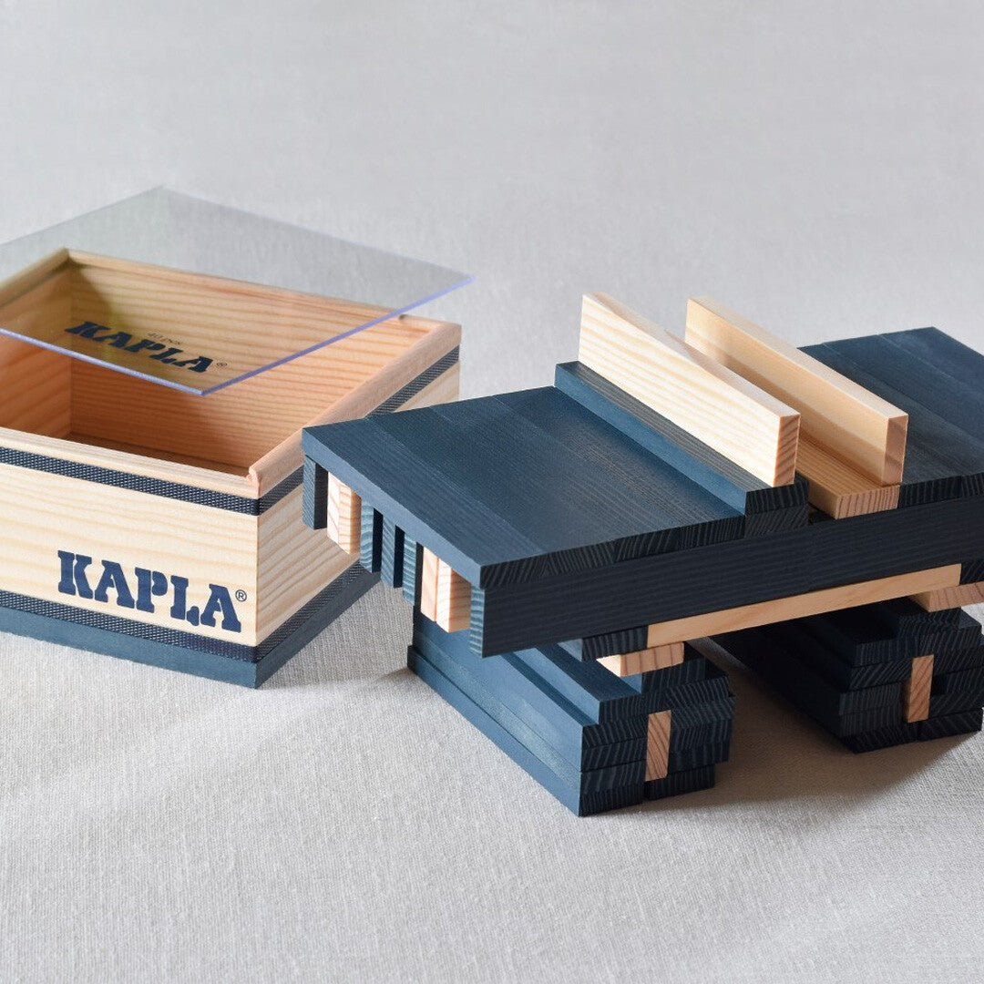 Een bouwwerk van Kapla houten blokken in donkerblauw en naturelkleur, naast de open houten opbergdoos.