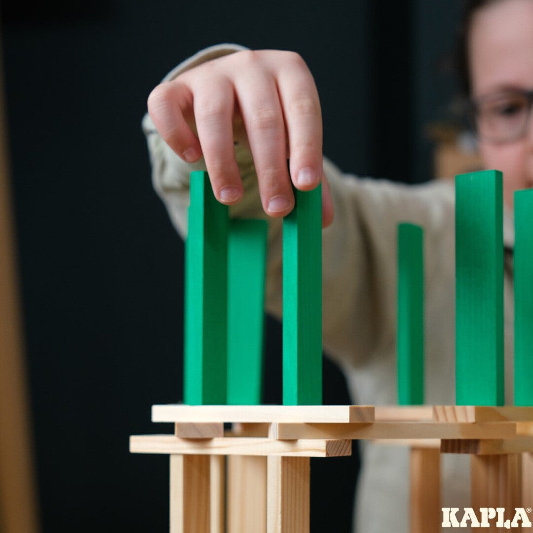 Een kinderhand plaatst een groen Kapla bouwblok op een houten constructie.